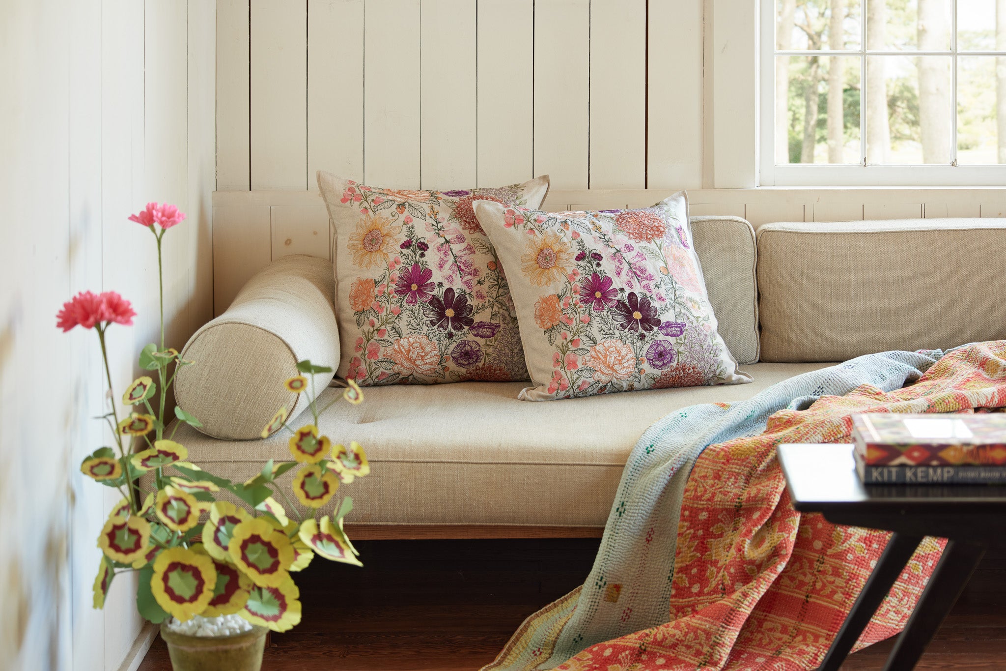 Couch with Coral and Tusk embroidered botanical Paradise Garden Pillows, The Green Vase paper geranium plant, kantha quilt throw and coffee table in front.