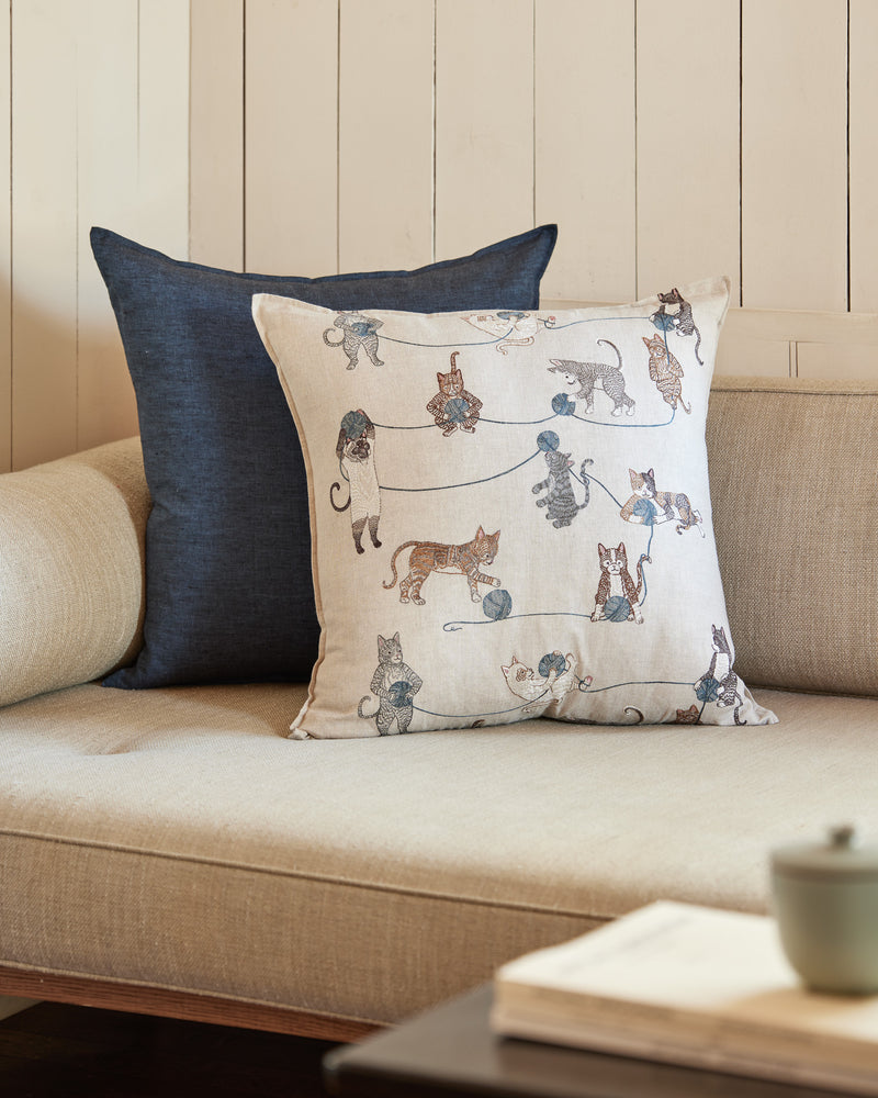 Corner of couch with dark blue pillow and Coral and Tusk embroidered Playful Cats pillow.