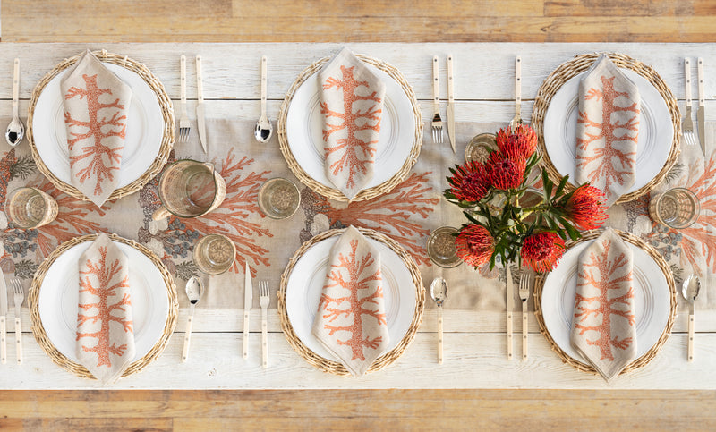 Overhead of dining table with six table settings, coastal themed, featuring Coral and Tusk embroidered coral motif table runner and dinner napkins, white plates on rattan placemats.