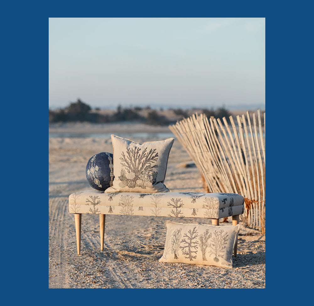 Coral and Tusk coastal coral motif embroidered pillows and bench on beach, coastal landscape in the background.