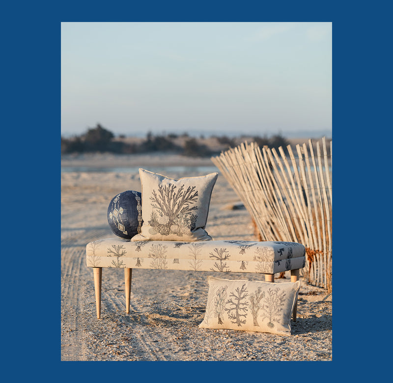 Coral and Tusk coastal coral motif embroidered pillows and bench on beach, coastal landscape in the background.