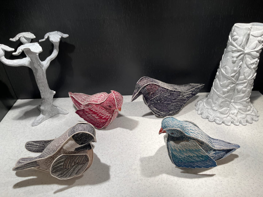 Four Coral & Tusk embroidered bird figurines in various colors (red, blue, gray, and black) arranged on a light gray surface against a dark background beside white ceramic candlestick holder and vase.