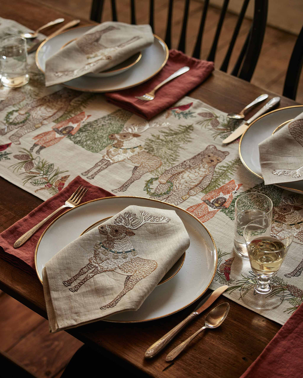 Coral & Tusk embroidered linen Christmas Cheer Table Runner and Christmas Cheer Reindeer Dinner Napkins styled on a table with silverware, glasses, red napkins, and ceramic plates.