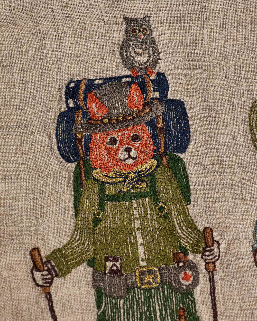 A close-up of Coral & Tusk Explorers Pouch, showing embroidery of Fox dressed as a camper, with a blue backpack, grey hat, green shirt and pants, gold scarf, map and first aid kit, and hiking sticks in hand, and an owl perched on top of the backpack.