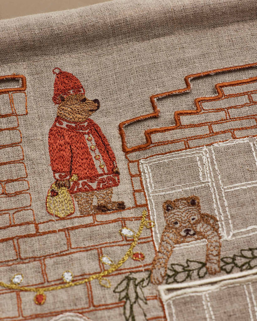 Close-up of embroidered Coral & Tusk Holiday House Advent Calendar featuring a brick house facade with multiple windows and hidden pockets, showing detail of Santa Bear standing on the rooftop holding a gold bag, a bear hanging a garland over a window, and Christmas lights strung along the roof.