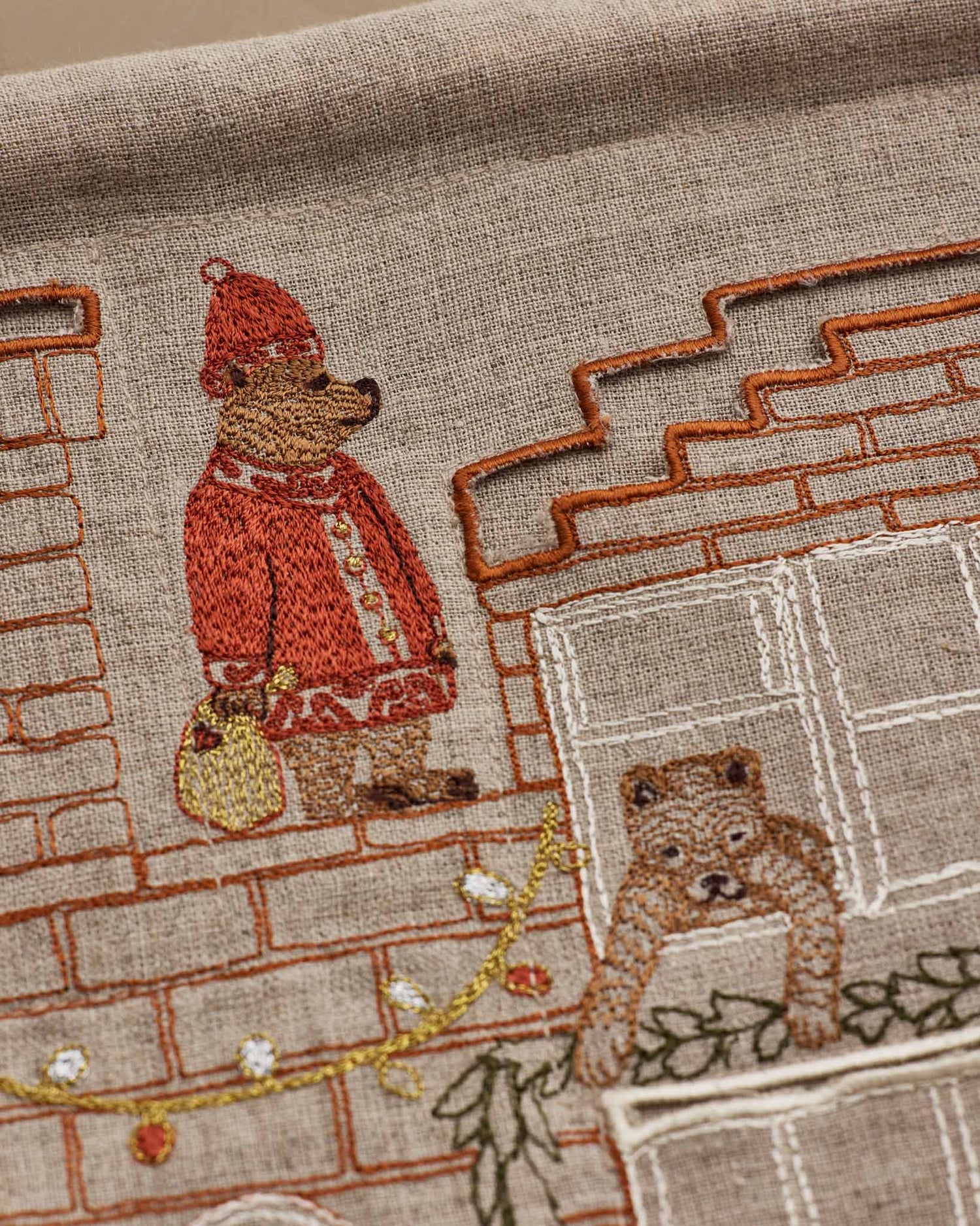 Close-up of embroidered Coral & Tusk Holiday House Advent Calendar featuring a brick house facade with multiple windows and hidden pockets, showing detail of Santa Bear standing on the rooftop holding a gold bag, a bear hanging a garland over a window, and Christmas lights strung along the roof.