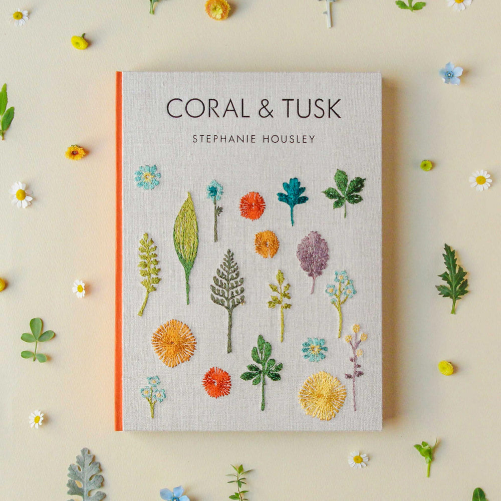 Book titled 'Coral & Tusk' by Stephanie Housley with floral embroidery on a yellow background styled with flowers and sprigs all around.