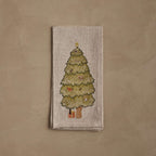 Coral & Tusk Peek a Tree Dinner Napkin, folded, featuring an embroidered Christmas tree with a gold star topper, hiding owl, bunny, squirrels, fox, and bear faces among the branches, with a wrapped orange gift at the base and an animal paw reaching down to grab it.