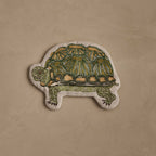 Coral & Tusk Turtle Pouch, turtle-shaped and featuring an embroidered tortoise in olive and dark greens, mustard yellow, and brown, on natural linen, zipper placement at the bottom of the shell.
