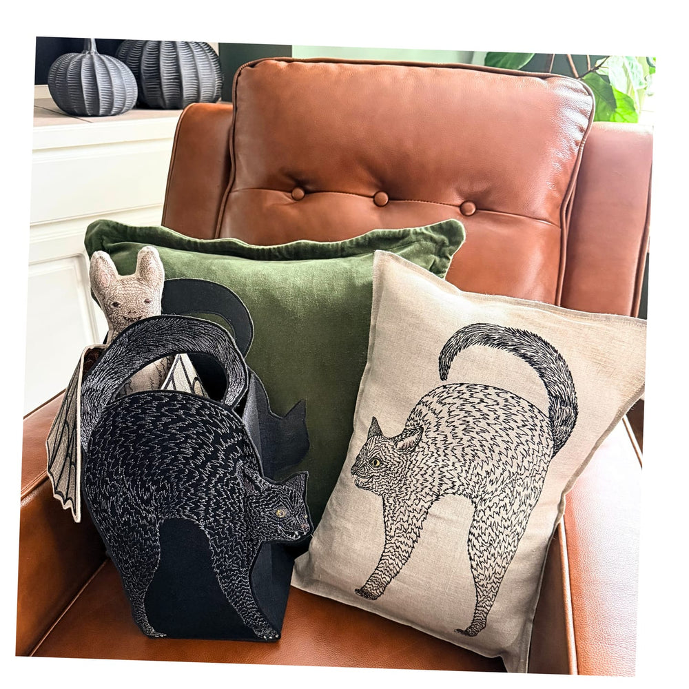 Two decorative throw pillows arranged on a brown leather armchair in a styled interior: the Coral & Tusk Scaredy Black Cat Pillow featuring a black cat silhouette, and a solid olive green pillow, with the Coral & Tusk Scaredy Cat Bag beside them and a Bat Doll peeking out.
