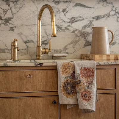 Coral & Tusk embroidered Autumn Botanical Tea Towels hanging on kitchen sink with marble backsplash, wooden cabinets, and gold faucet.