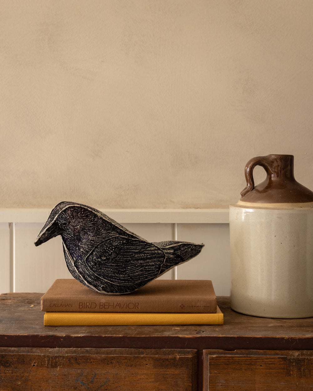 Coral & Tusk Blackbird of Mystery decorative bird resting on two stacked books on a rustic wooden table, beside a large ceramic jug against a beige wall.