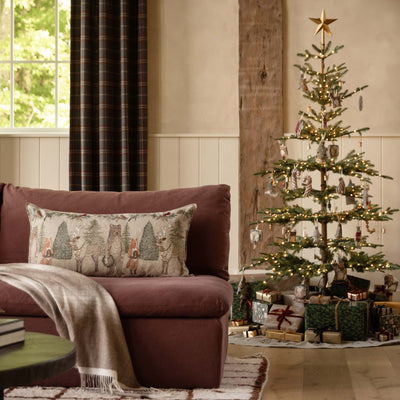 A cozy living room scene with a maroon sofa, a beige throw blanket, and a Coral & Tusk Christmas Cheers decorative pillow featuring a woodland animal embroidery. To the right stands a lit Christmas tree with ornaments and wrapped gifts underneath, set in a warm interior with curtains, a wood wall panel, and a window.