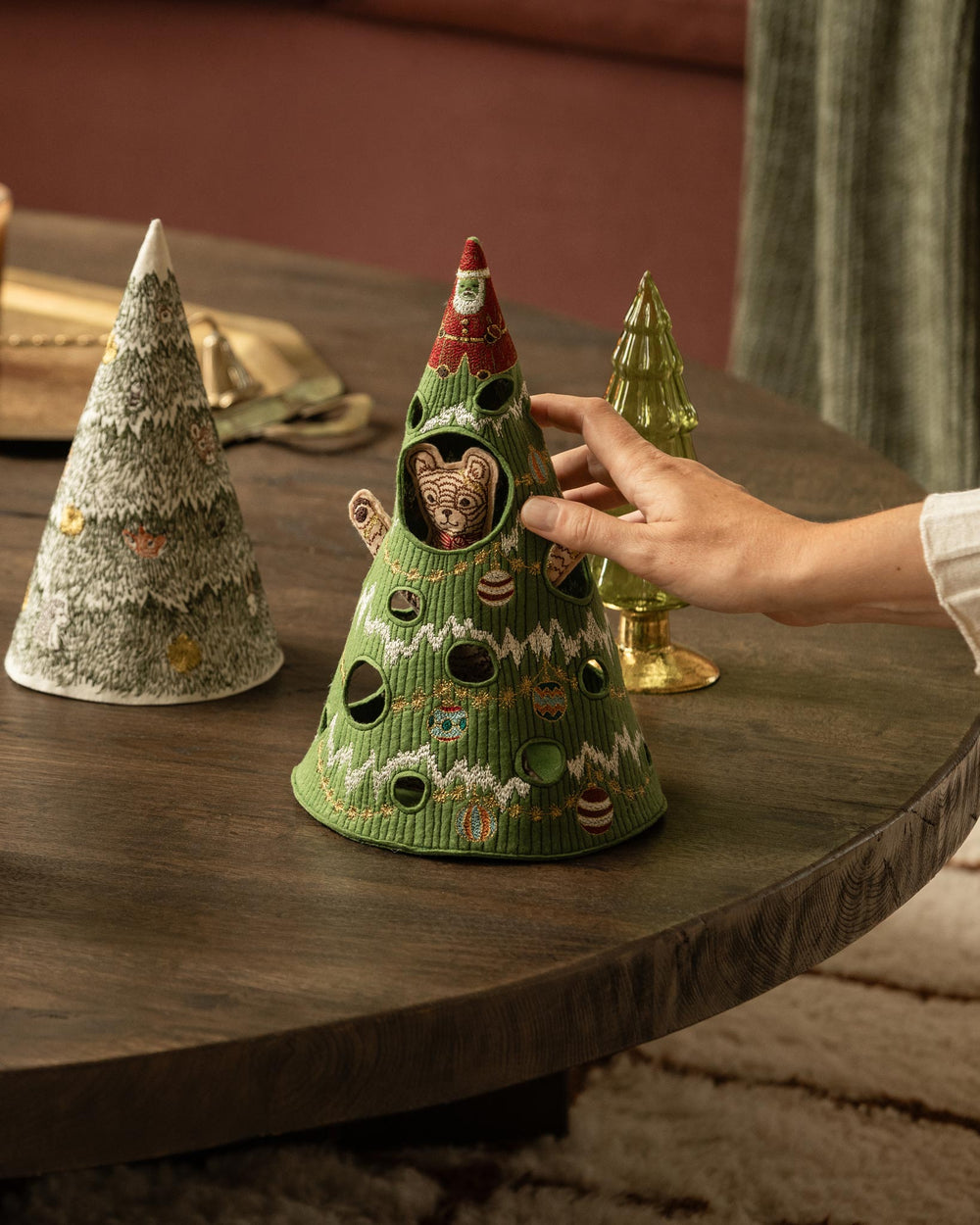 A hand holds the Coral & Tusk Christmas Tree Bear, a Christmas tree-shaped embroidered felt object with a bear peeking out from the center, set on a dark wooden table next to two other decorative trees.