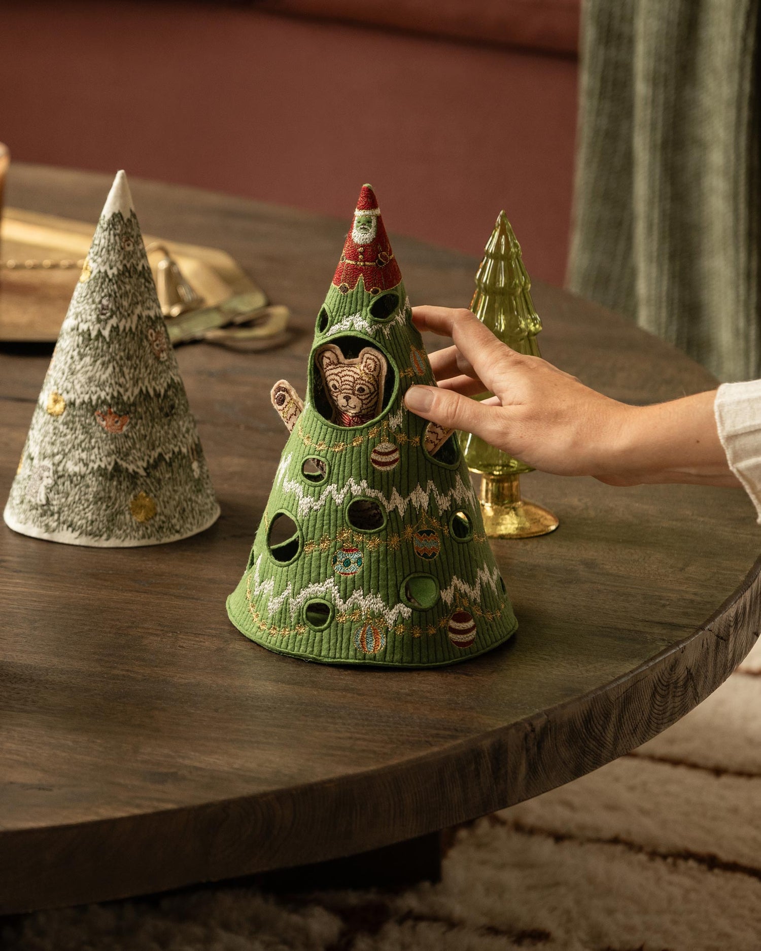 A hand holds the Coral & Tusk Christmas Tree Bear, a Christmas tree-shaped embroidered felt object with a bear peeking out from the center, set on a dark wooden table next to two other decorative trees.