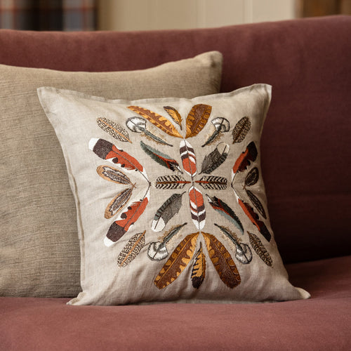 Coral & Tusk Feather Radiance throw pillow, featuring an intricately embroidered circular radiating feather pattern, layered in front of a textured plain neutral beige pillow resting on a maroon sofa.