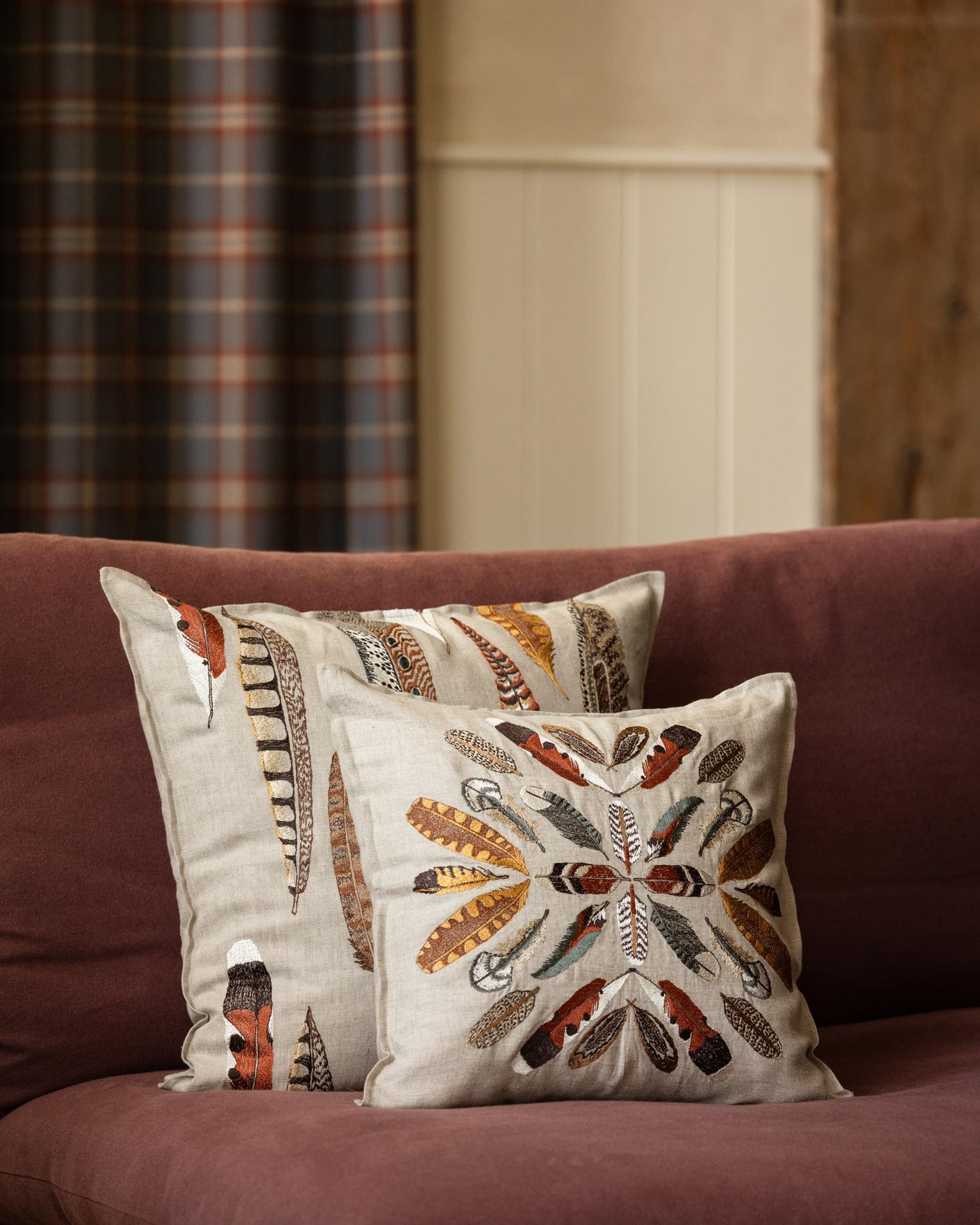 A styled living room scene featuring a maroon sectional sofa with four Coral & Tusk feather-themed decorative embroidered throw pillows, a side table with a lamp, a Coral & Tusk Feather Tissue Box Cover, and a coffee mug, and a round coffee table in the foreground with books.