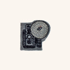The Fox Camera Accessory is a small gray felt item featuring dark charcoal embroidery of a vintage twin-lens reflex camera with a circular metallic silver thread coil representing a flash attachment to the right of the lenses.