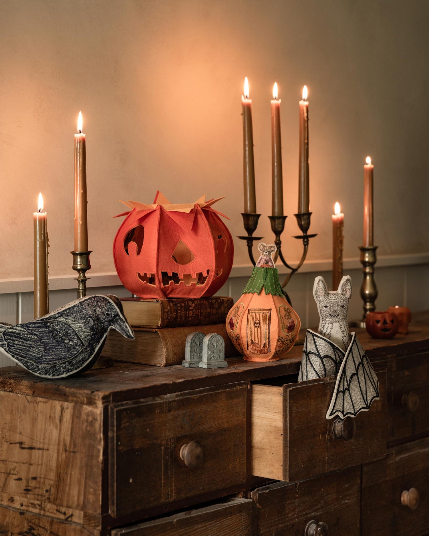 A styled Halloween decoration scene with Coral & Tusk decorative objects, including embroidered pumpkins, a jack-o'-lantern trick-or-treat bag, a blackbird, a bat, and lit taper candles, on a wooden dresser against a beige wall.