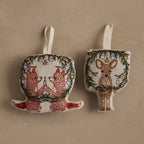 Coral & Tusk Togetherness Ornament Set, two embroidered linen ornaments: two foxes holding a candle inside an evergreen wreath with silver and gold ornaments, and Deer peering through a matching wreath, each with a cream hanging loop.