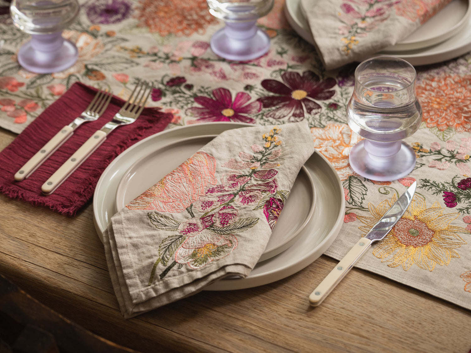 Coral & Tusk embroidered Paradise Garden Table Runner and Blush Bouquet Dinner Napkin, styled with cutlery and glasses on a wooden table.