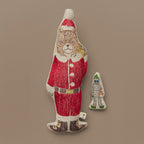The Coral & Tusk Santa Pocket Doll is an embroidered linen doll stuffed with polyfill. The brown bear doll is displayed flat against a beige wall, and wears a red and white Santa suit and hat, along with a black belt and shoes, both with gold accents. Over one shoulder the Santa Bear carries a gold bag for delivering gifts. The yeti pal is grey with blue eyes, carrying a brown teddy bear. They are wearing a green elf hat and shoes, and can be placed on the back of the Santa Bear Doll in the gold bag pocket.