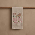 Coral & Tusk embroidered linen Stockings Tea Towel, folded and hanging on a wood rod, depicting a brown and white kitten and brown puppy each hanging in red and white striped stockings. The stockings have holy berry sprigs at the tops and green heels.