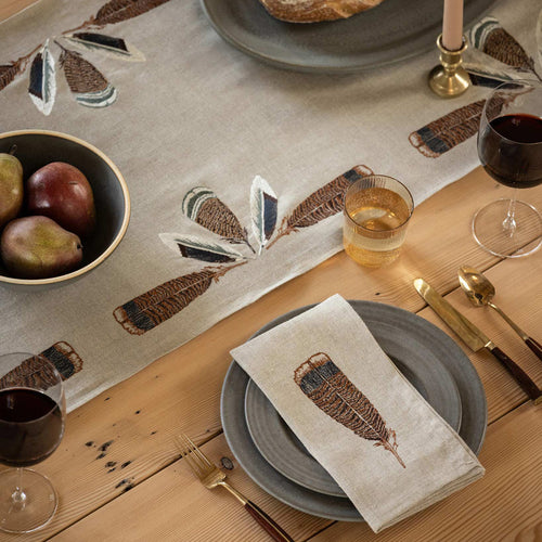 A single place setting on a wooden dining table, centered around the Coral & Tusk Turkey Feather Border Table Runner, with a slate gray ceramic plate, Coral & Tusk Turkey Feather Dinner Napkin, glass, silverware, a serving plate with a loaf of bread, and a bowl of pears.