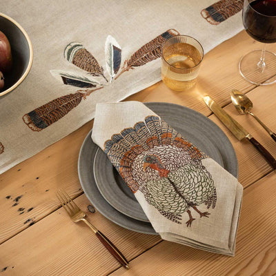 Overhead view of a dining table showing one table setting featuring Coral & Tusk embroidered linen Turkey Feather Border Table Runner in the center, slate gray ceramic plates with Coral & Tusk Wild Turkey Dinner Napkin, glasses, silverware, a serving plate with a loaf of bread, and a bowl of pears on a wooden table.