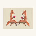 The Seconds Sale Smitten Foxes Card features an embroidered illustration of two orange foxes sitting on a wooden bench facing each other, each holding a small pink heart in its front paws on light cream handmade paper.