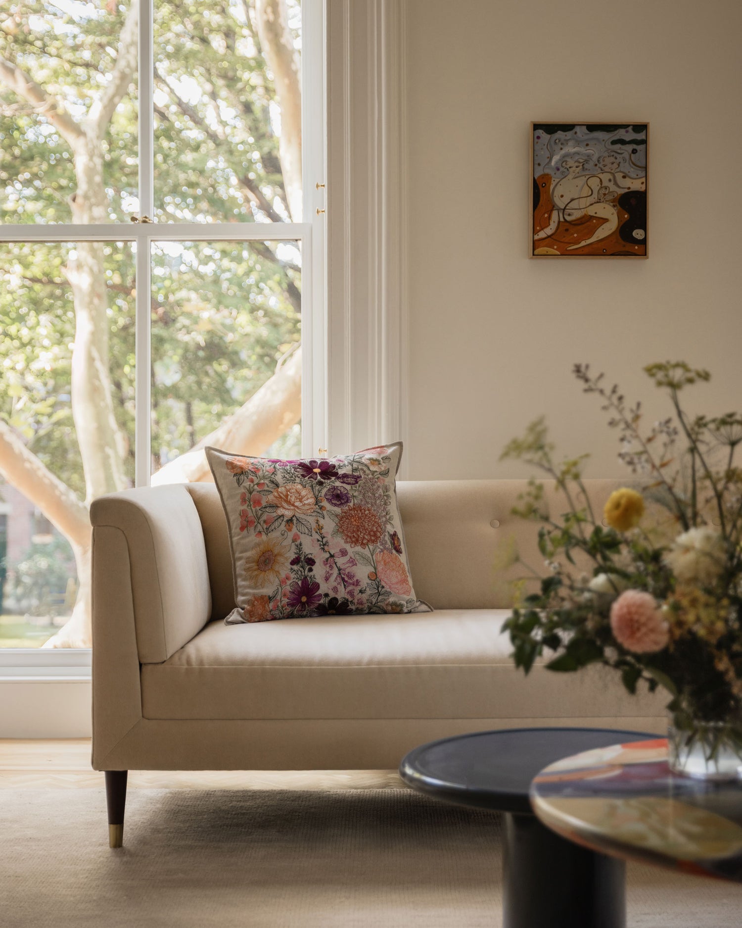 Coral & Tusk Paradise Garden Pillow on a white sofa in a living room with large windows and a vase of flowers on a table.