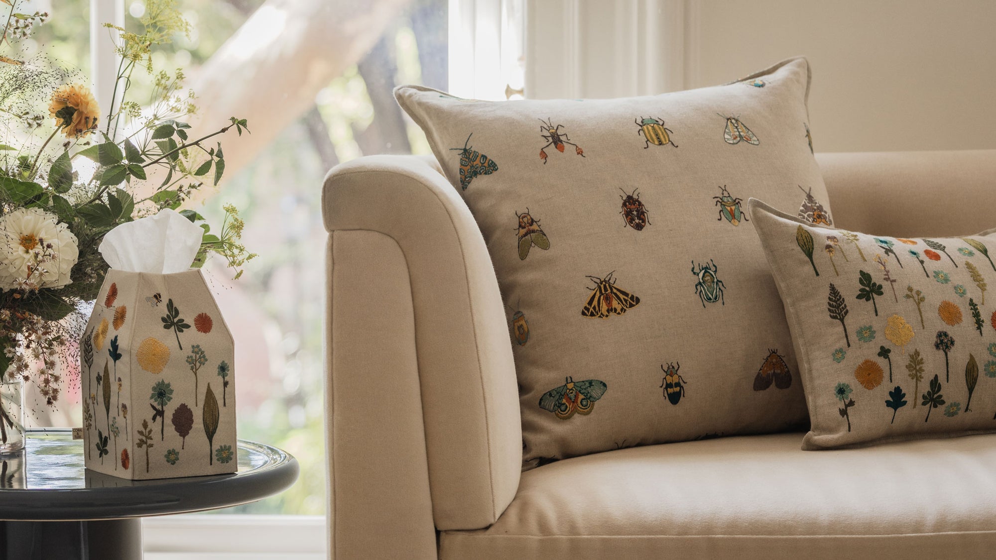 Off-white sofa with a Coral & Tusk embroidered Wings Pillow and Garden Pillow, next to a side table holding a bouquet of flowers and Coral & Tusk Garden Tissue Box Cover, in front of a window with bright natural light.