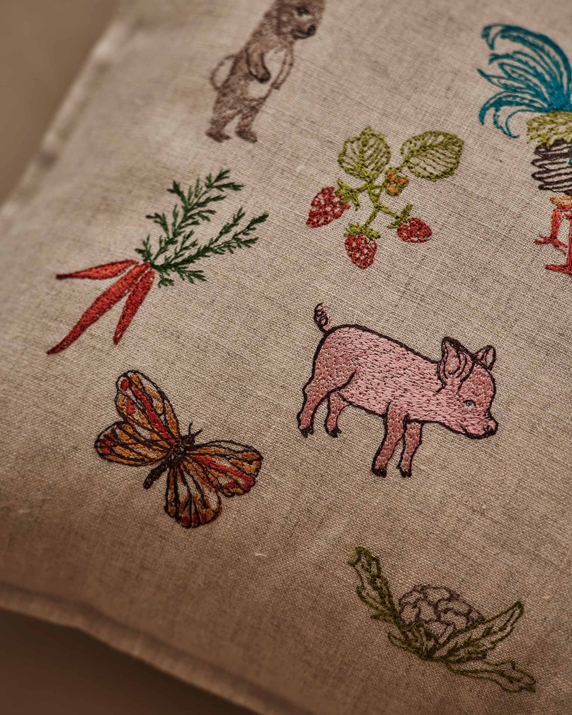 Close-up of Coral & Tusk 12 × 16-inch embroidered linen Garden Gathering Pillow depicting farm and garden life, featuring a pig, strawberries, carrots, cauliflower, and a butterfly.