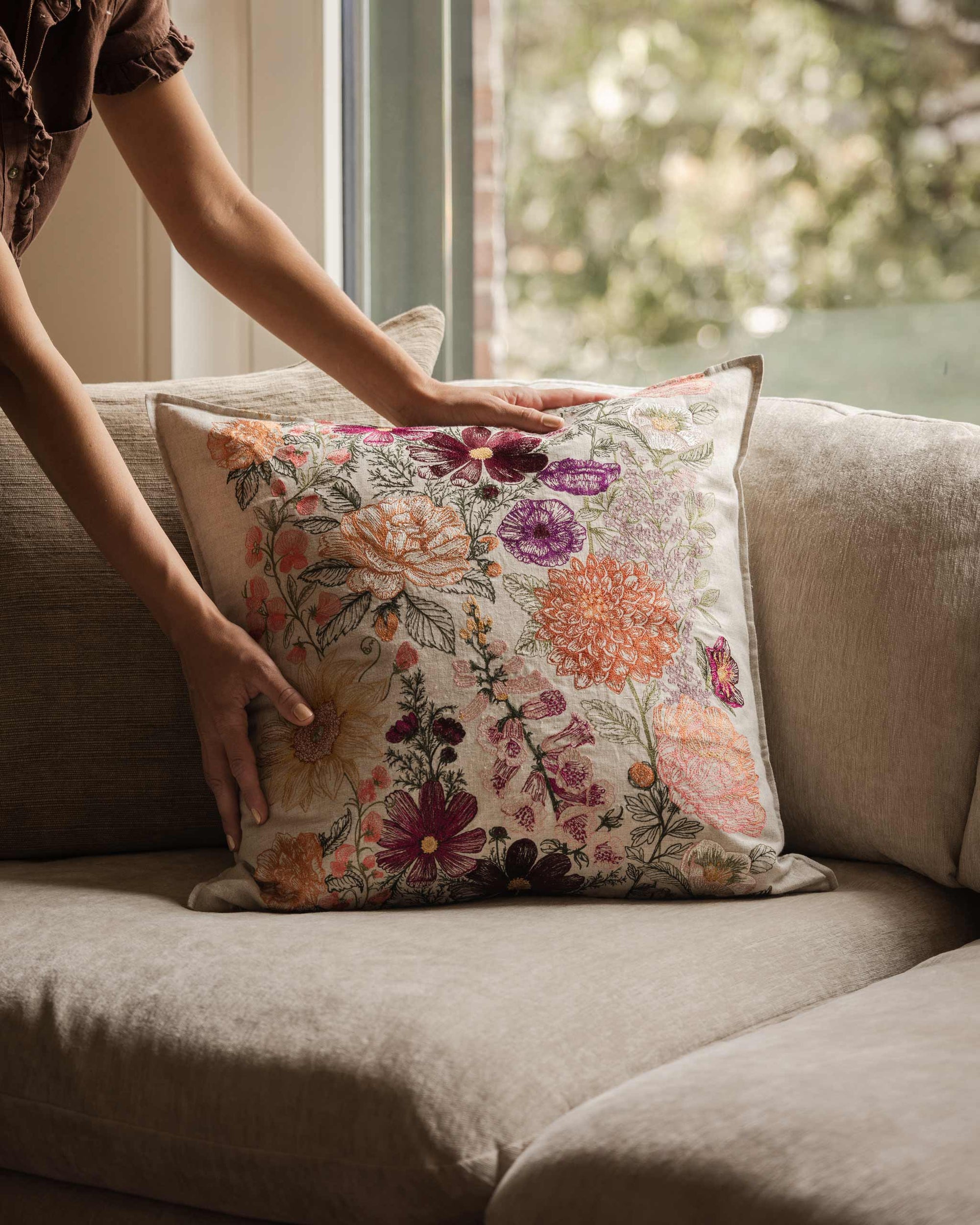 Person arranging a Coral & Tusk embroidered Paradise Garden Pillow on a couch near a window with soft natural light and a view of greenery.