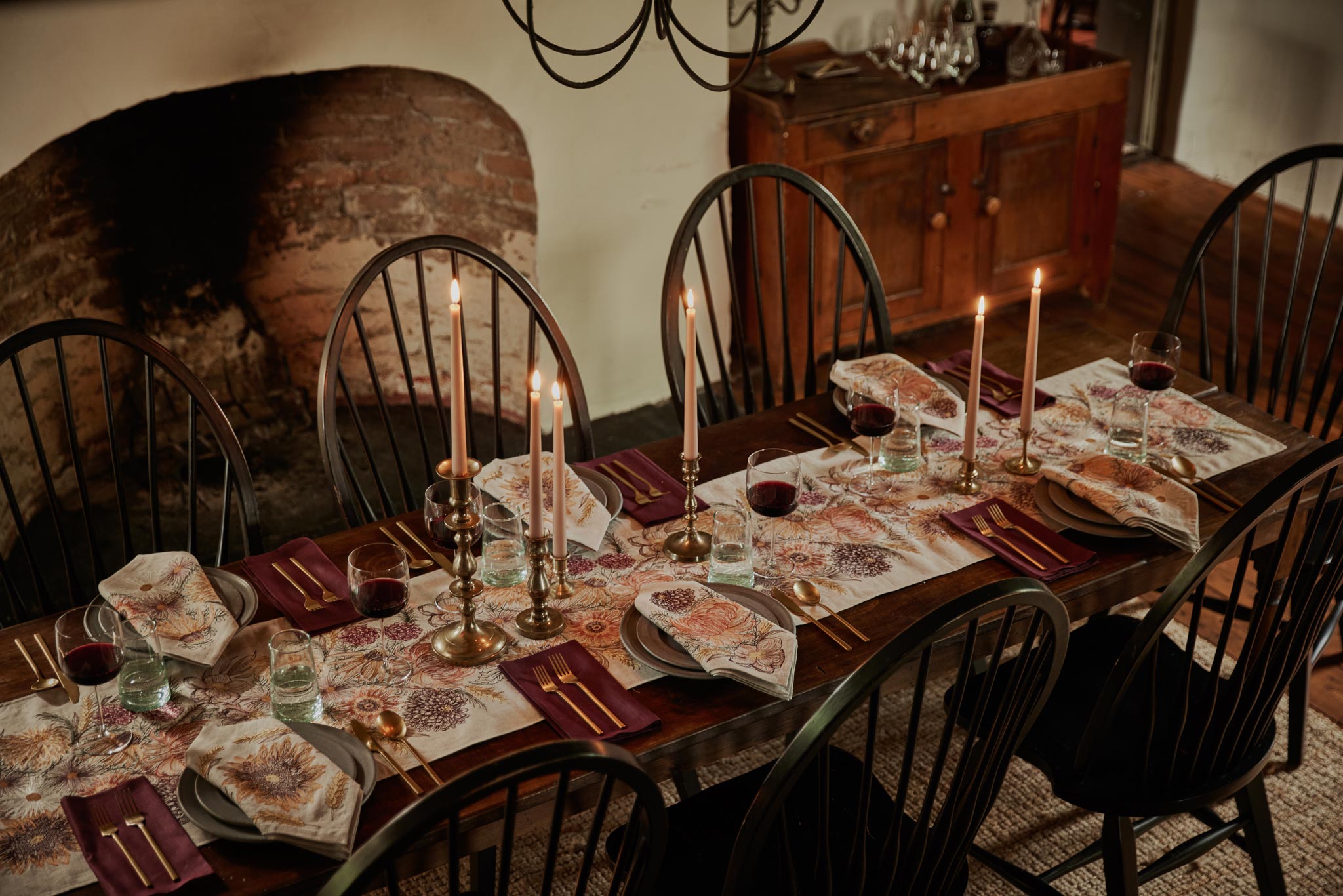 Coral & Tusk embroidered linen Autumn Blossoms Table Runner and Autumn Blossoms Dinner Napkins styled in a dining room on a table in front of a fireplace with six place settings, brass silverware, lit candles, wine glasses, and gray ceramic plates.