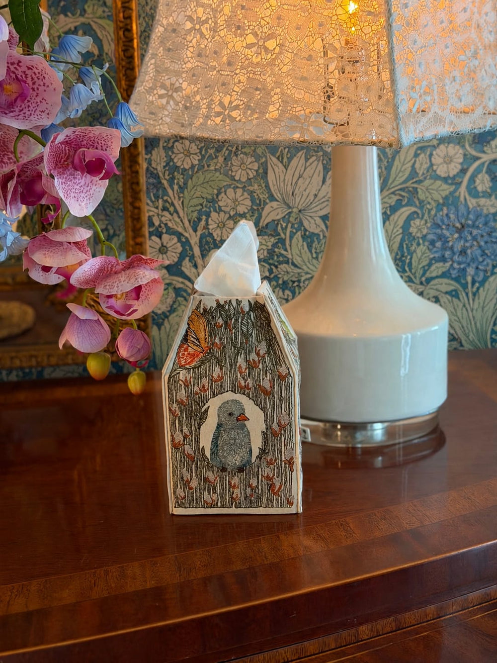 A Coral & Tusk decorative Birdhouse tissue box cover shaped like a birdhouse, with a bluebird in the center, sits on a wooden table beside a lit table lamp, with pink orchids and floral wallpaper in the background.