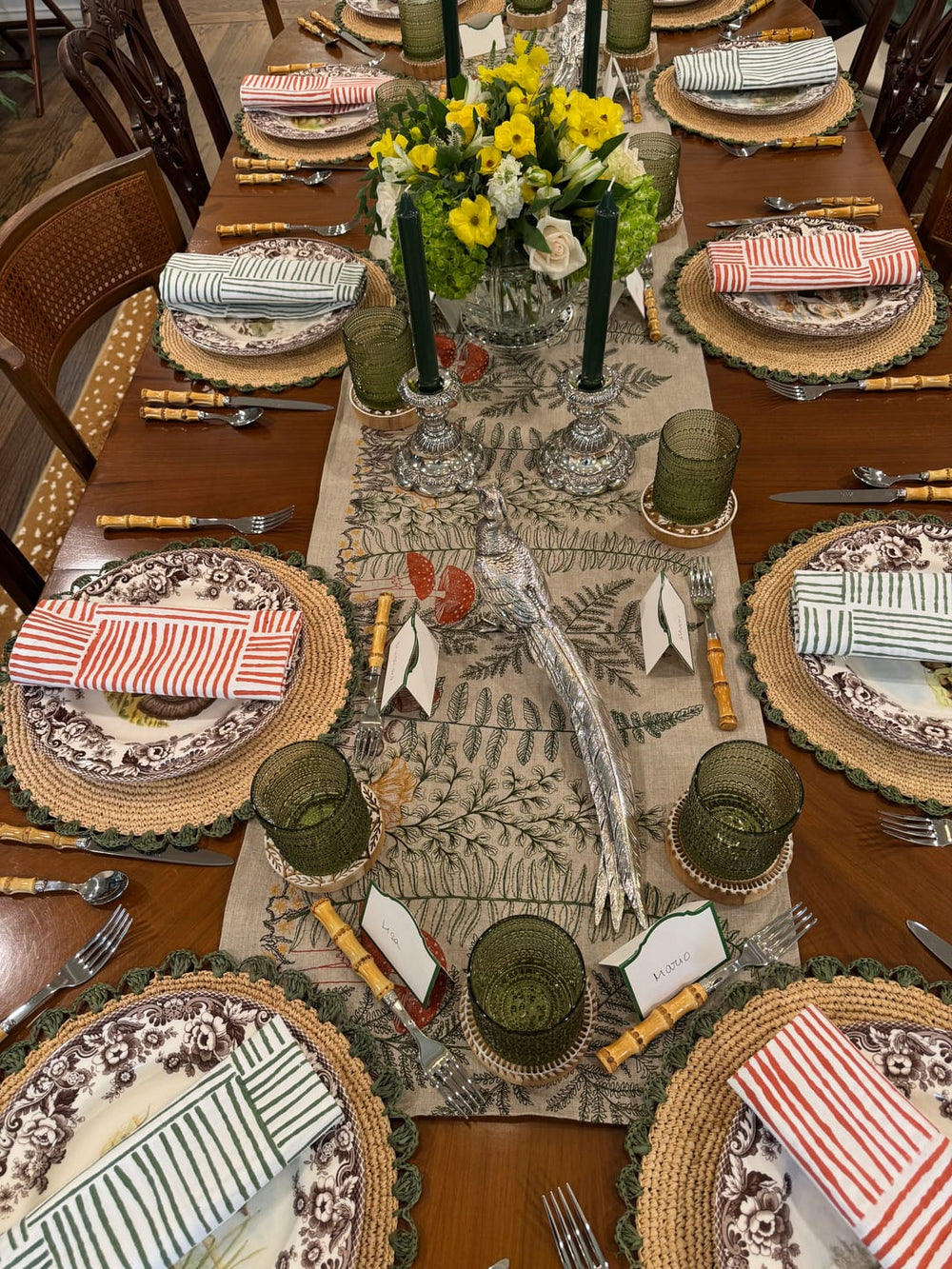 A fully set dining table for eight, featuring brown and white floral patterned plates, woven placemats, striped napkins, green glassware, and a Coral & Tusk Mushrooms and Ferns botanical runner with a central floral arrangement.